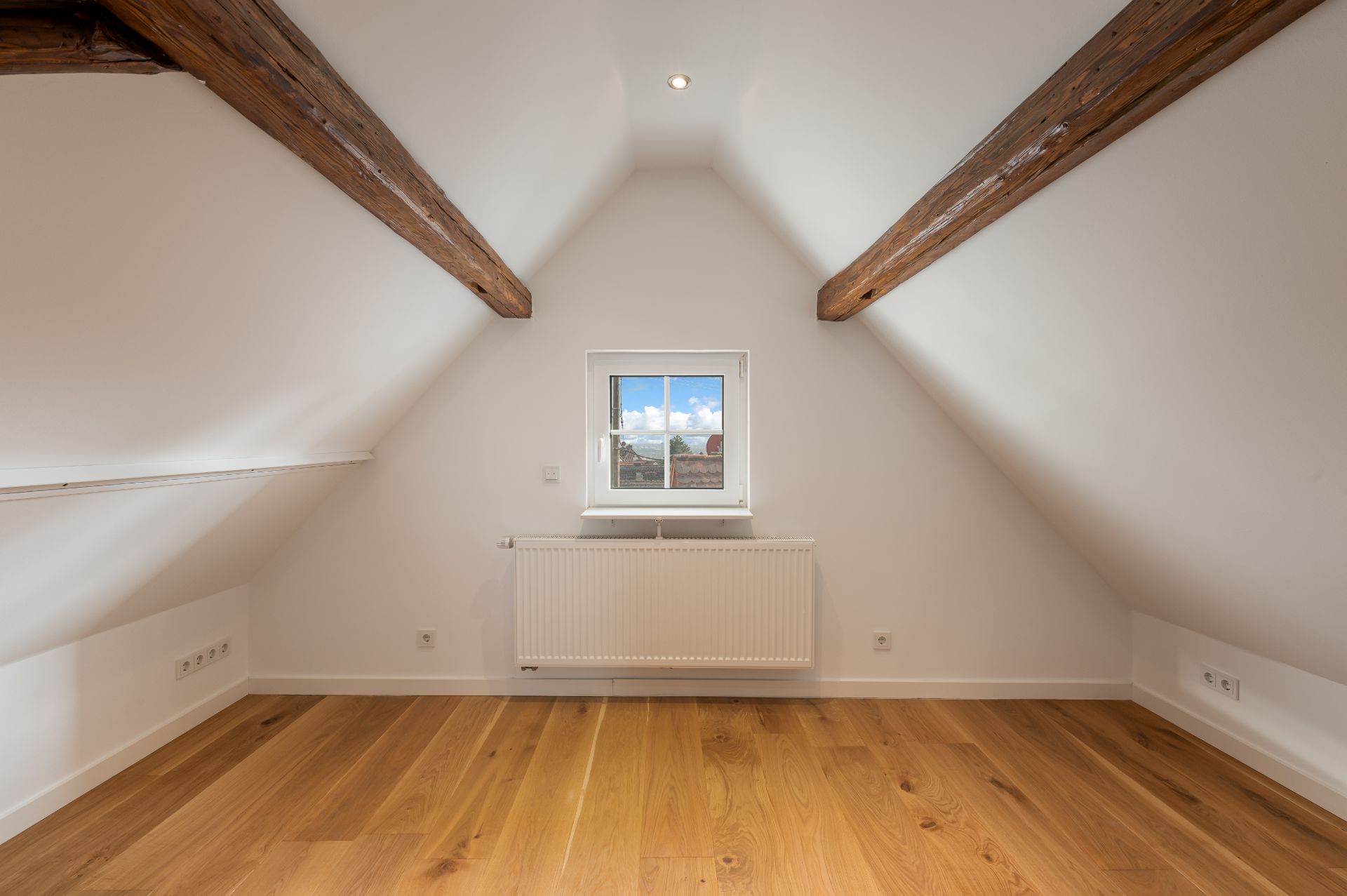 Leerer Dachraum mit Holzfußboden, sichtbaren Balken und kleinem Fenster mit Heizkörper darunter.