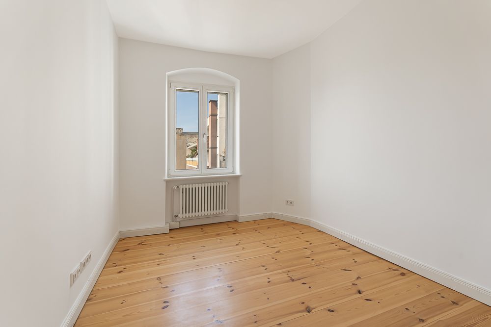 Leerer Raum mit Holzboden, weiß gestrichenen Wänden und Fenster mit Heizkörper darunter.