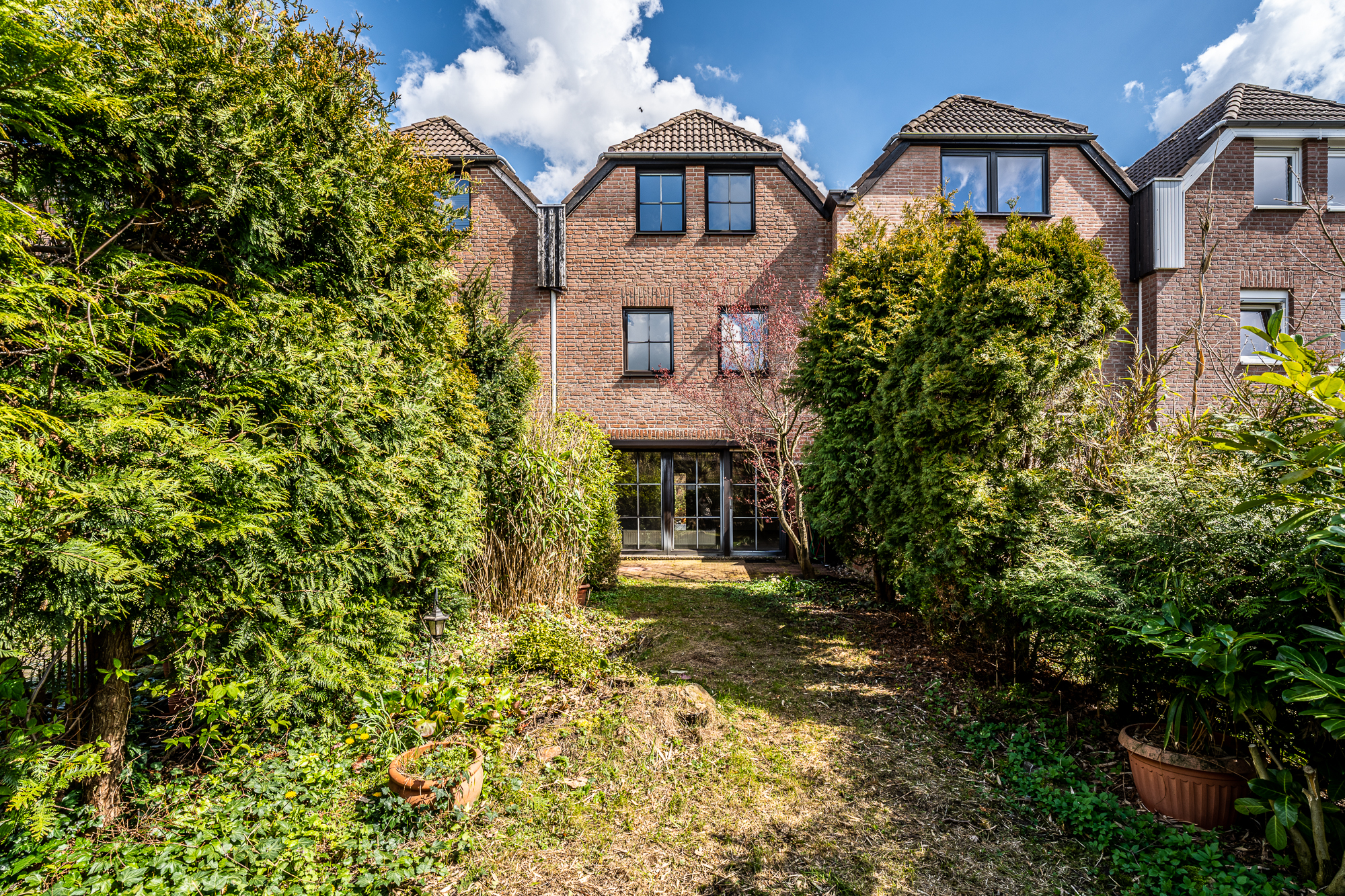 Rückansicht eines Backsteinhauses mit Garten und großen Büschen im Vordergrund.