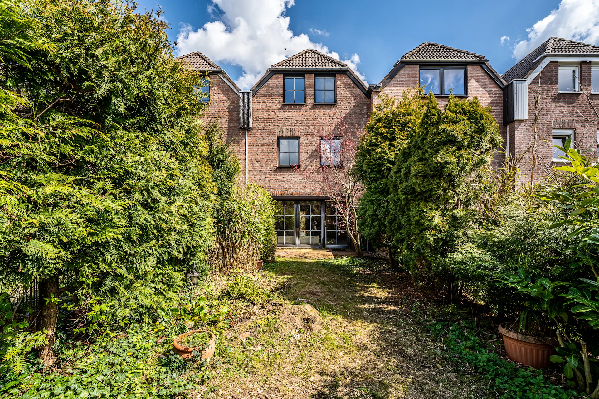 Rückansicht eines Backsteinhauses mit Garten und großen Büschen im Vordergrund.