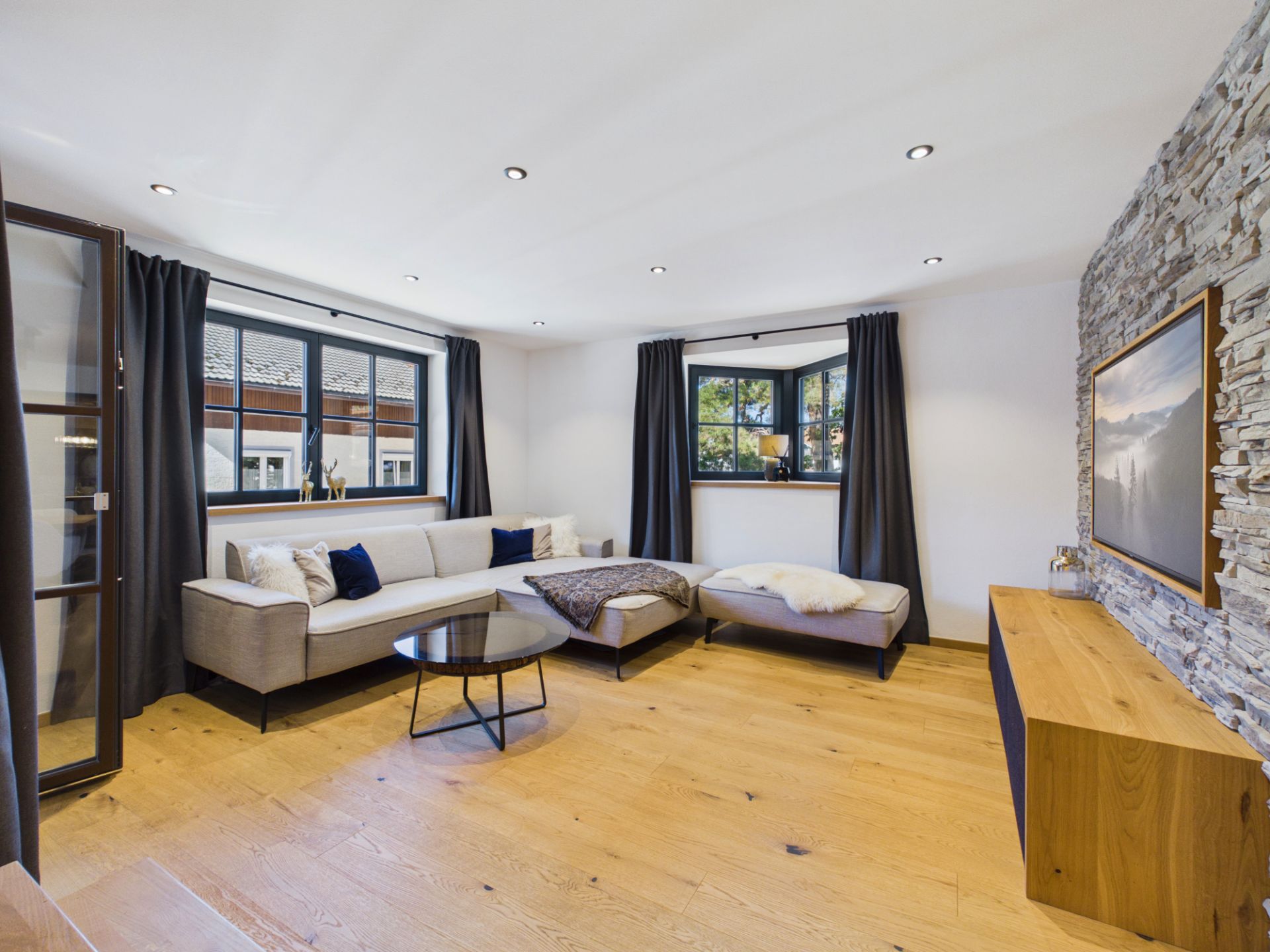 Modernes Wohnzimmer mit grauem Sofa, Holzfußboden, Steinwand und großen Fenstern mit Vorhängen.