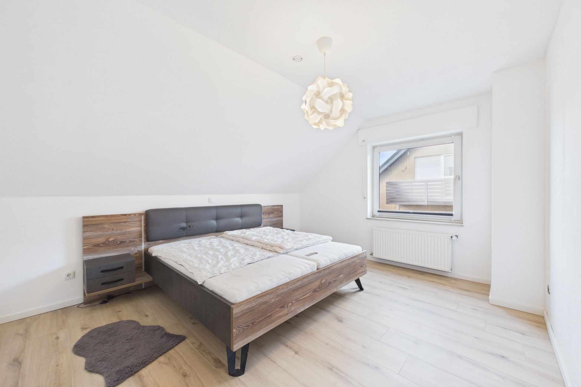 Helles Schlafzimmer mit Doppelbett, Nachttisch, Fenster und Holzfußboden.