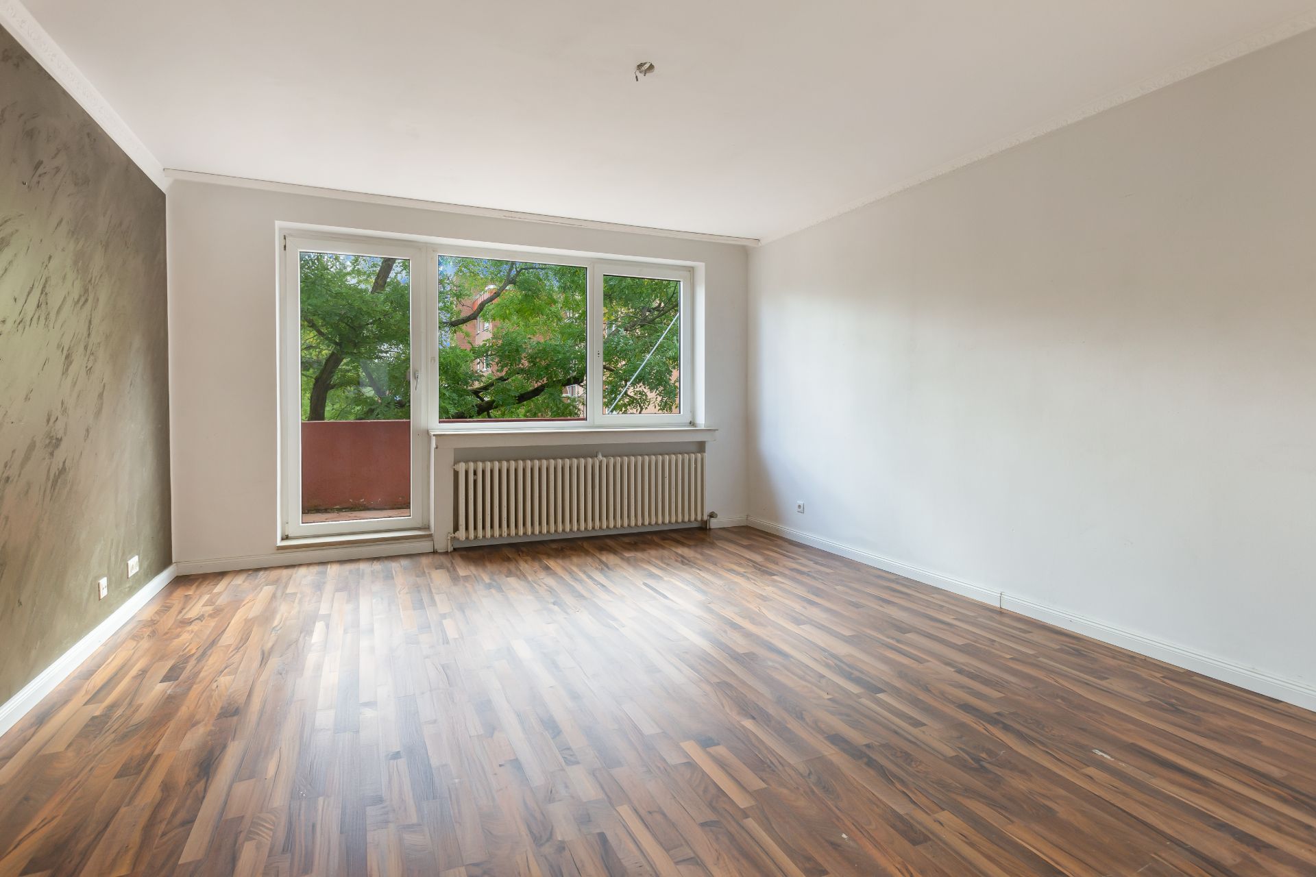 Heller leerer Raum mit Holzboden, großem Fenster und Heizkörper darunter.