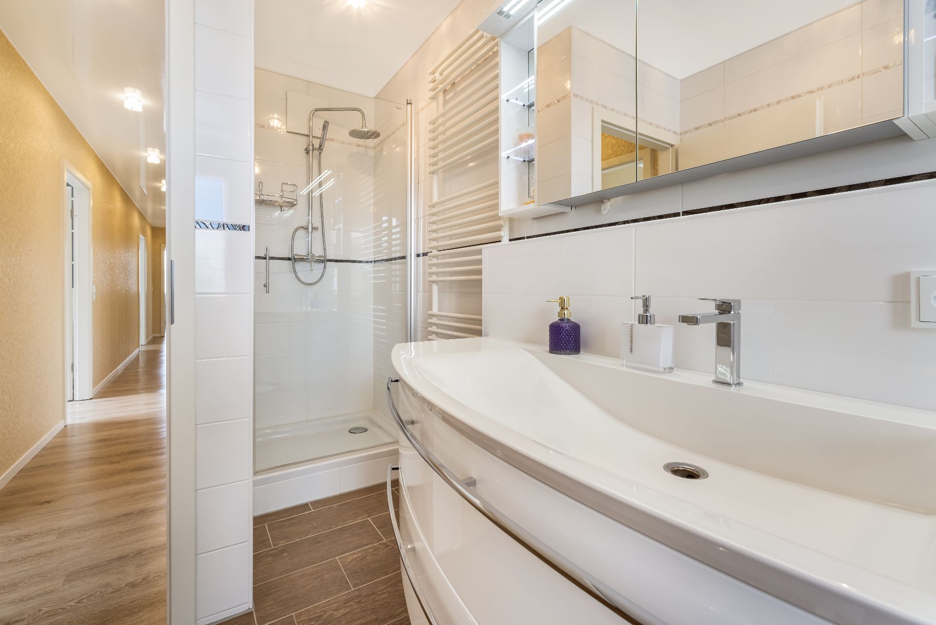 Modernes Badezimmer mit Dusche, Doppelwaschbecken und großem Spiegel, angrenzender Flur mit Holzboden.