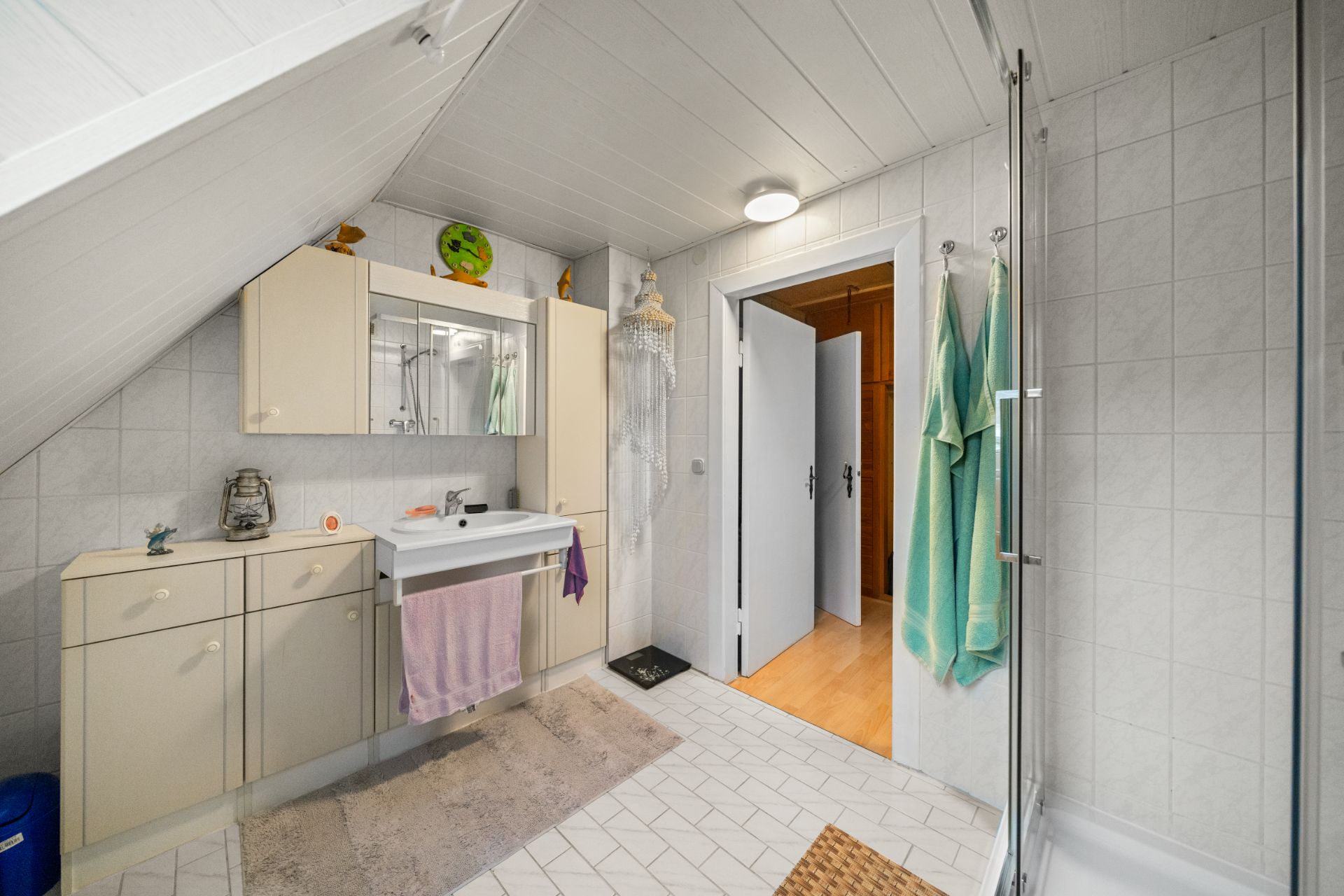 Helles Badezimmer mit Waschbecken, Spiegel, Hängeschrank und Dusche, weiße Fliesen und Holzdecke.