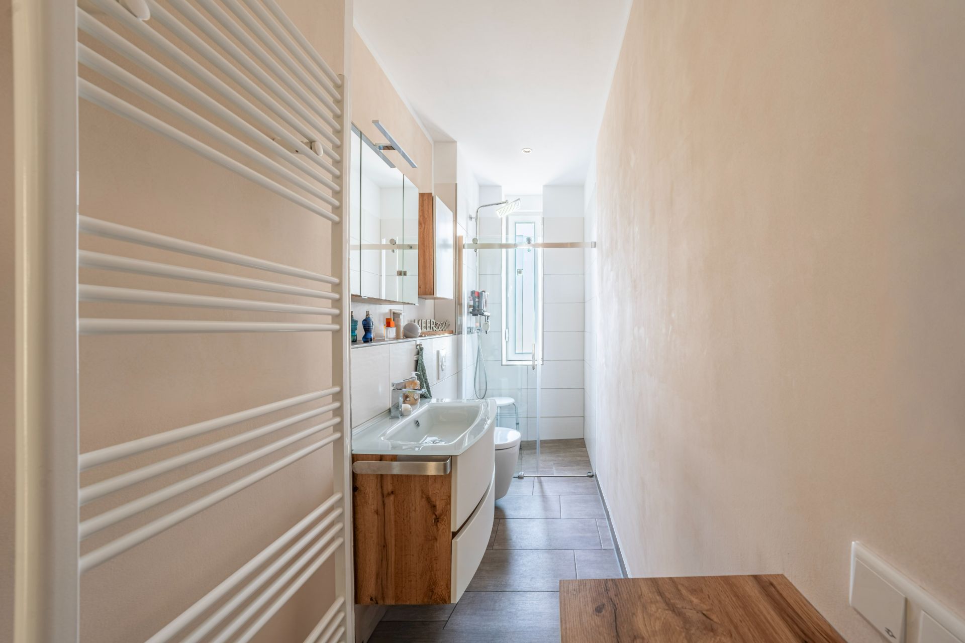 Helles, schmales Badezimmer mit Waschbecken, Spiegel, Dusche und Fenster am Ende.