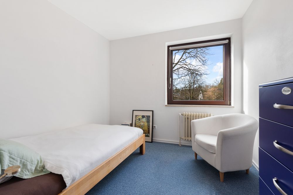 Ein schlicht eingerichtetes Zimmer mit Bett, Sessel, Kommode und Fenster mit Blick nach draußen.