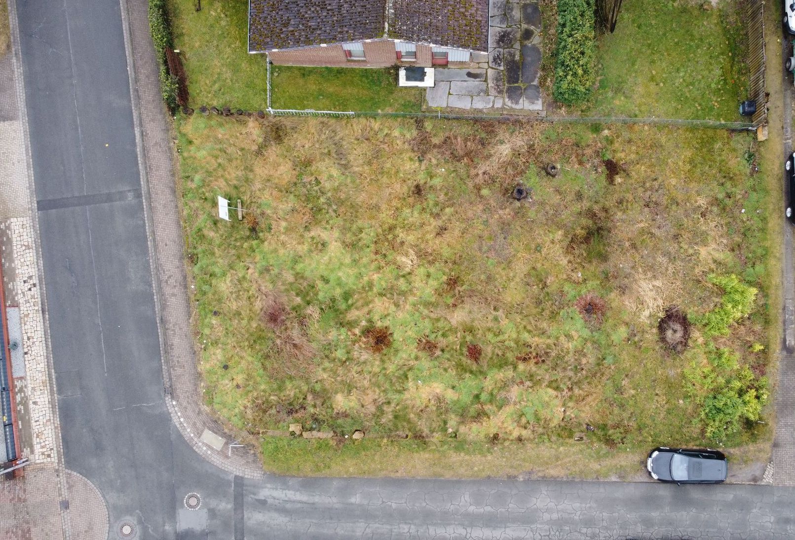Luftbild eines Grundstücks mit Grasfläche, daneben ein Haus und geparkte Autos an der Straße.