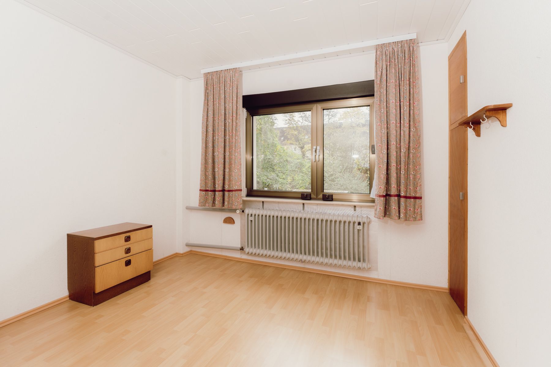 Leeres Zimmer mit Parkettboden, Fenster mit Vorhängen, Heizung und einer kleinen Kommode.