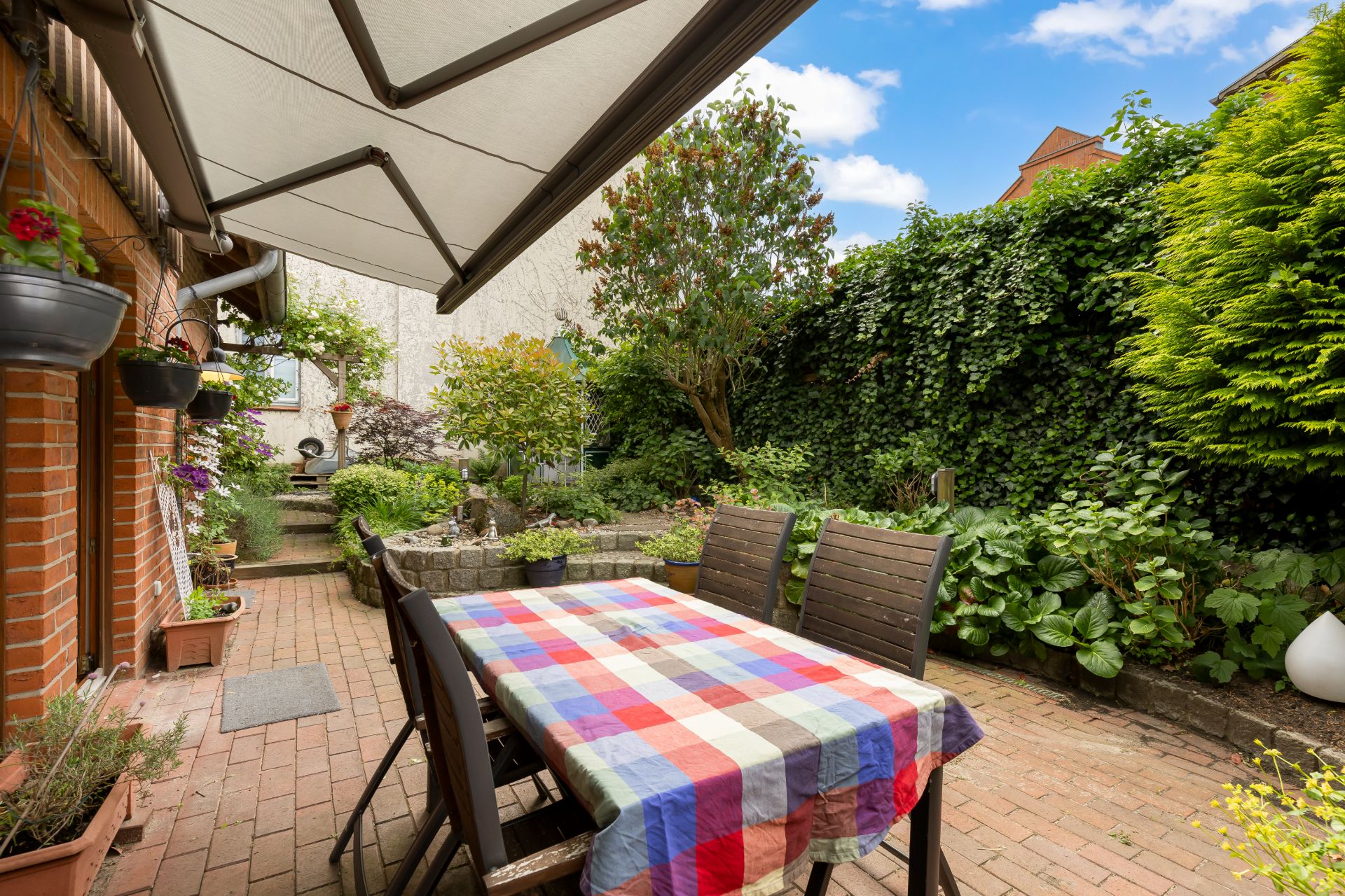 Gepflasterte Terrasse mit buntem Tisch, Stühlen und Garten mit vielen Pflanzen.