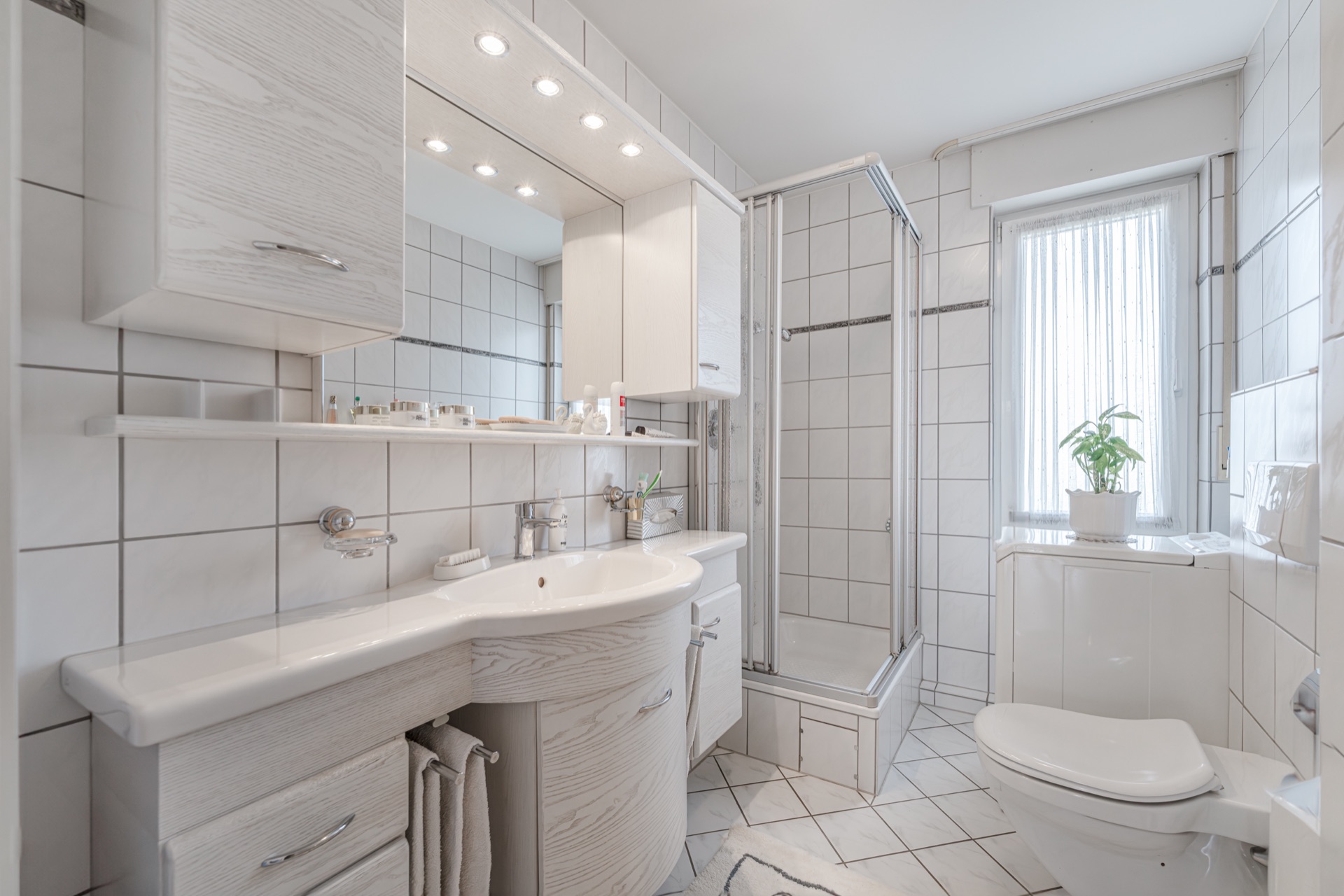 Modernes, weiß gefliestes Badezimmer mit Dusche, Waschbecken, WC und Fenster mit Pflanze.