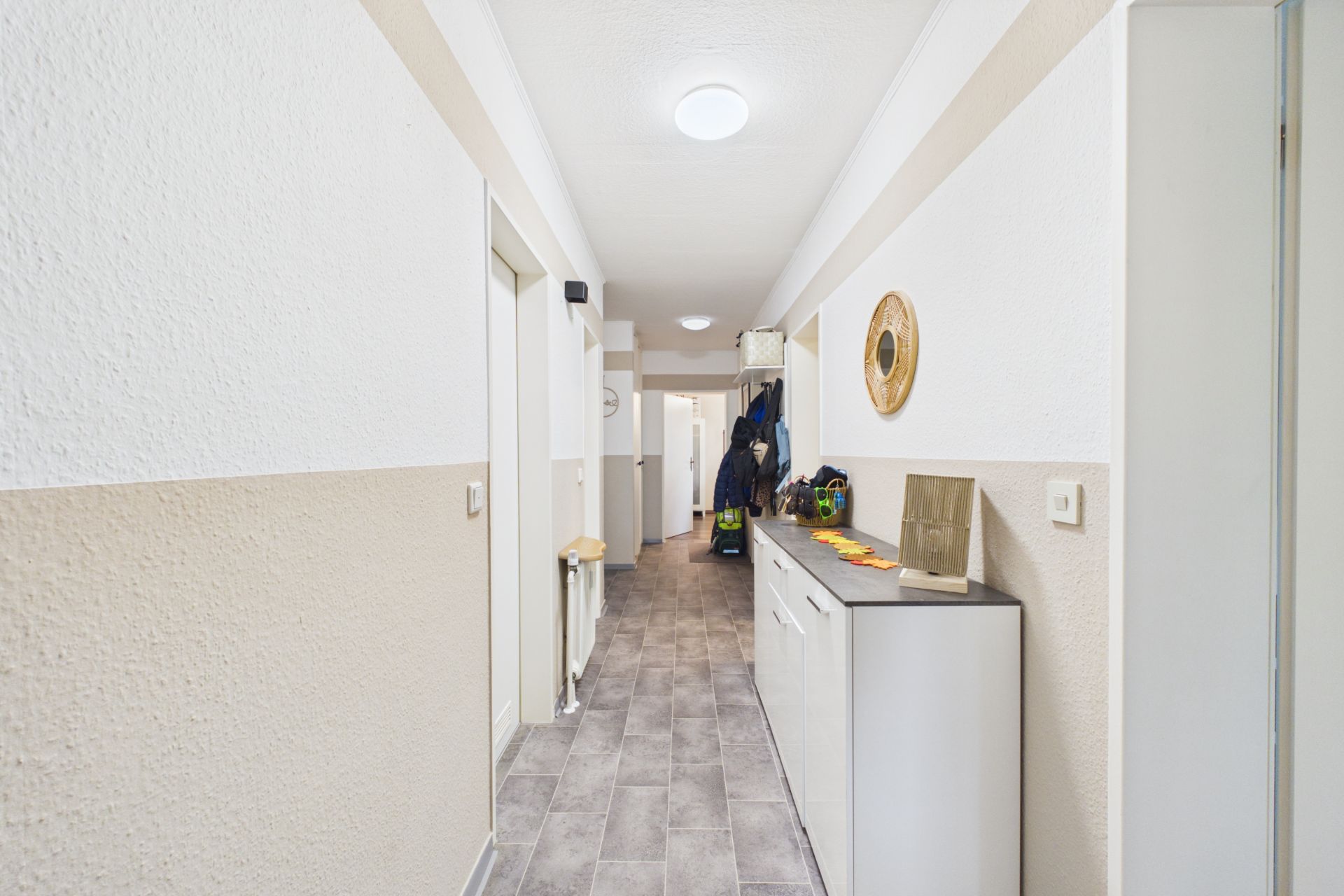 Langer Flur mit Fliesenboden, weißen Wänden, Kommode und Garderobe mit Jacken.