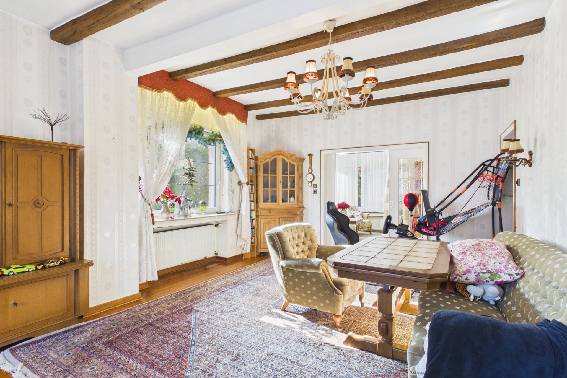 Wohnzimmer mit Teppich, Holzbalken, Essecke, Schaukelliege und großen Fenstern mit Gardinen.