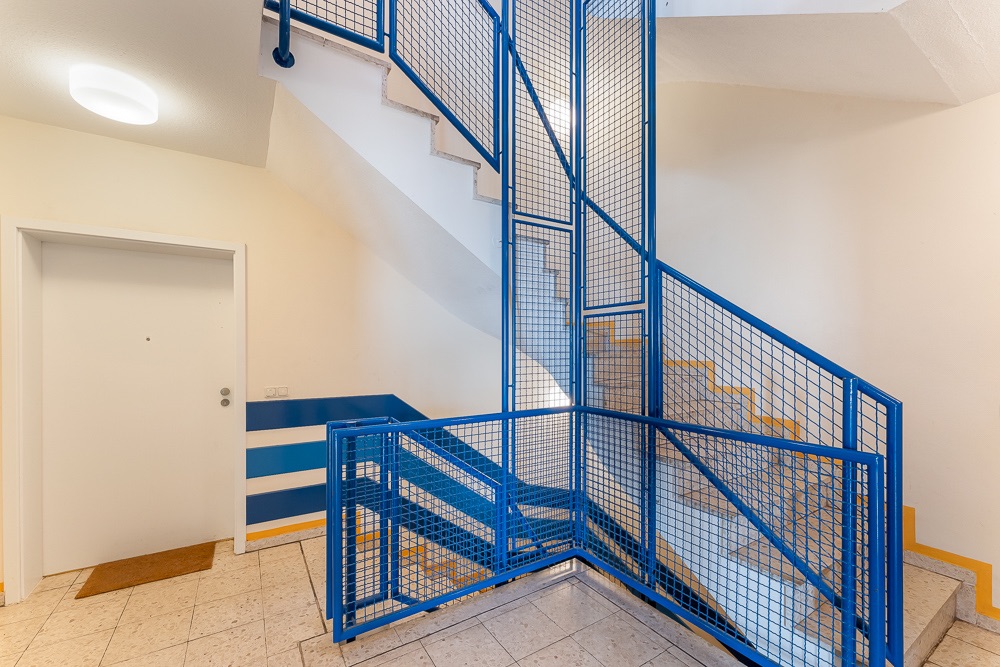 Treppenhaus mit blauem Geländer und Eingangstür auf einem Flur in einem Wohngebäude.
