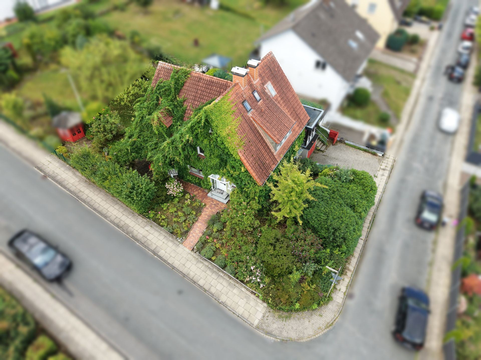 Luftaufnahme eines Wohnhauses mit rotem Dach und grünem Garten an einer Straßenecke.
