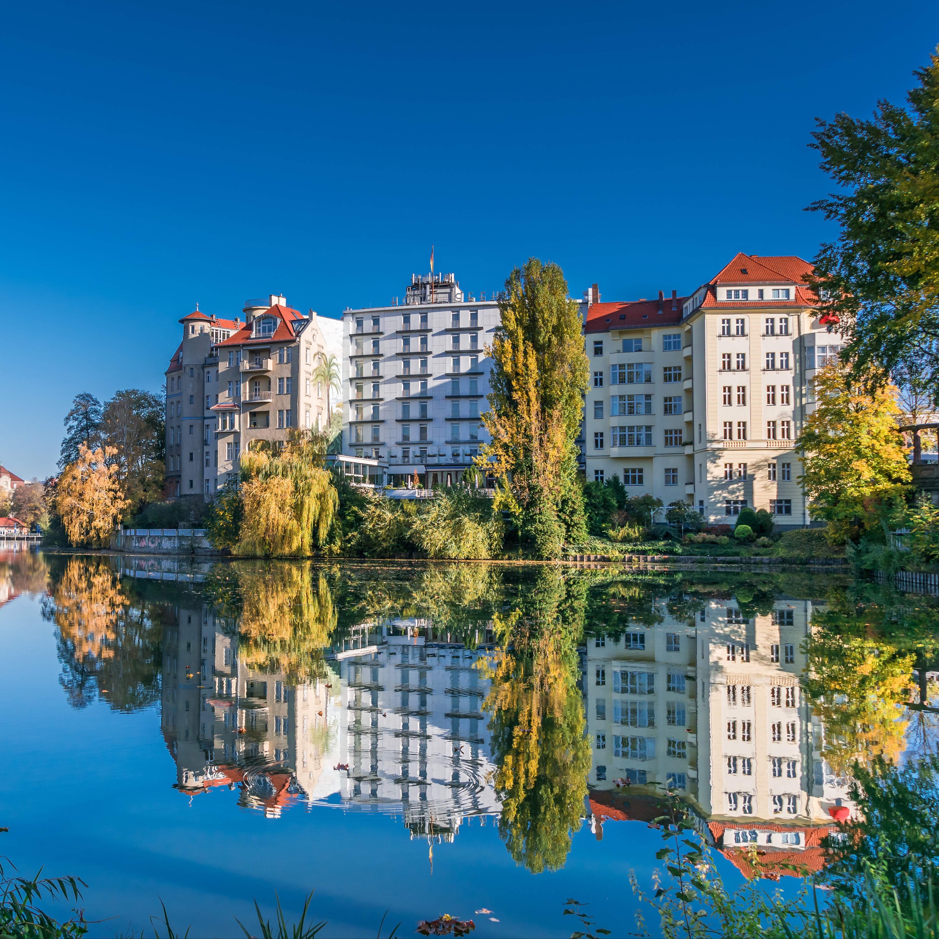 Mehrstöckige Wohnhäuser am Fluss mit Spiegelung im Wasser, umgeben von Bäumen.