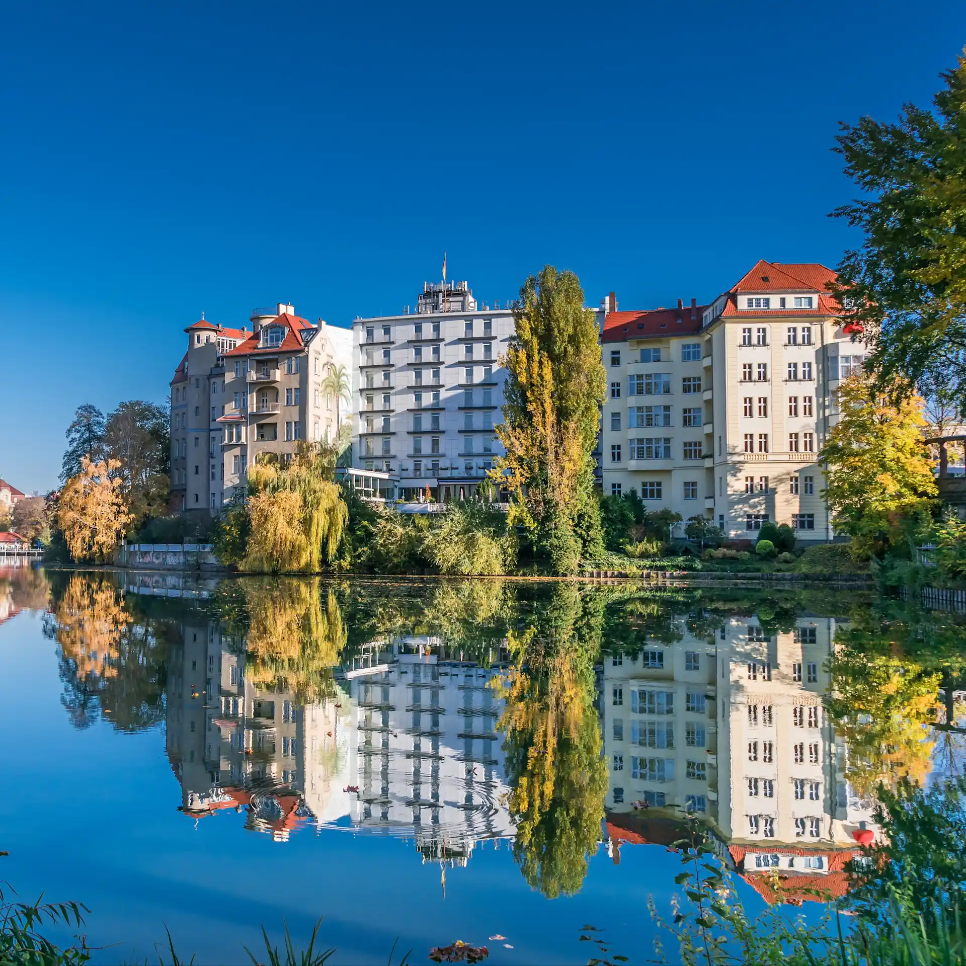 Mehrstöckige Wohnhäuser am Fluss mit Spiegelung im Wasser, umgeben von Bäumen.