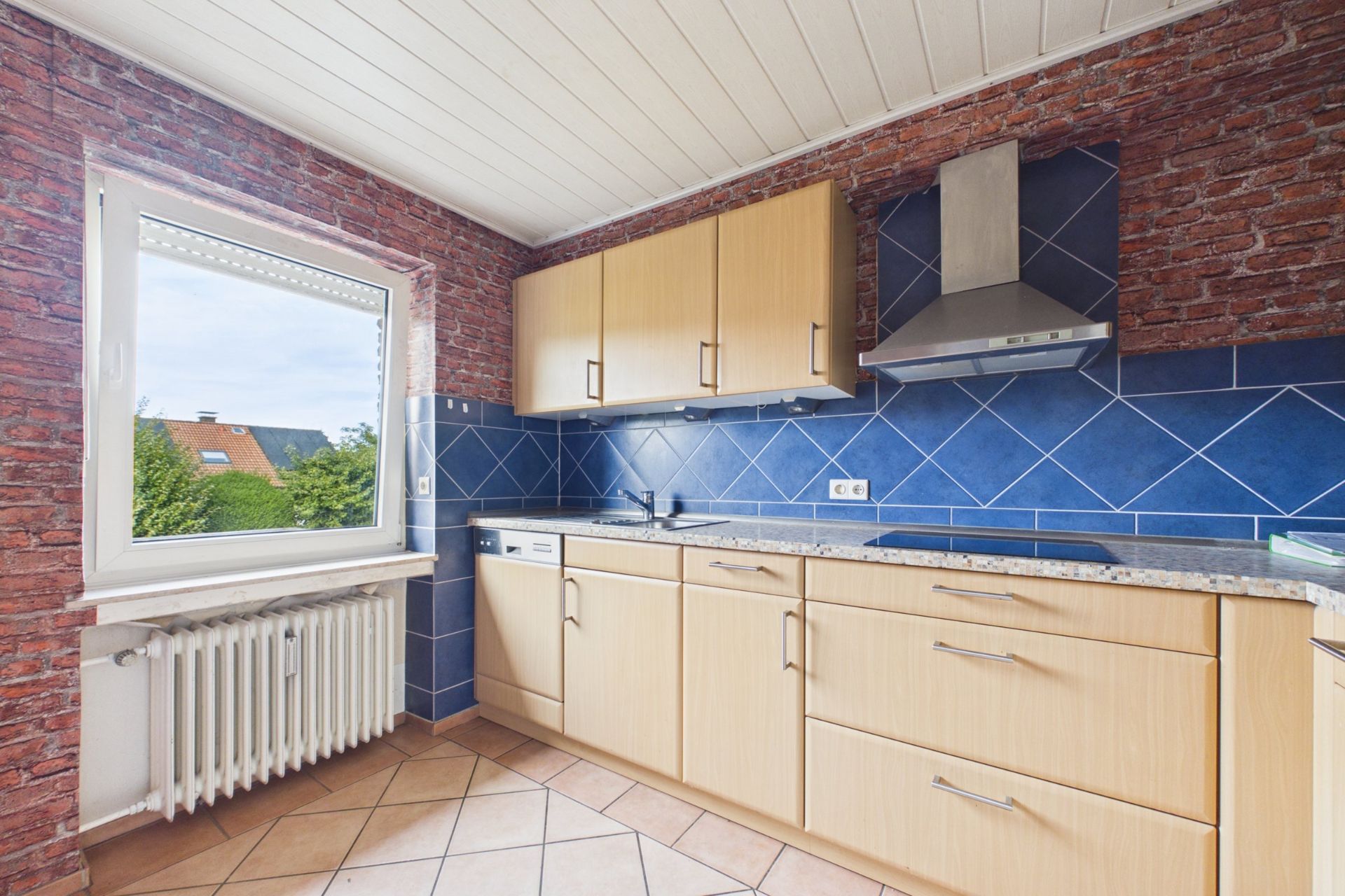 Helle Küche mit Holzschränken, blauem Fliesenspiegel und großem Fenster.
