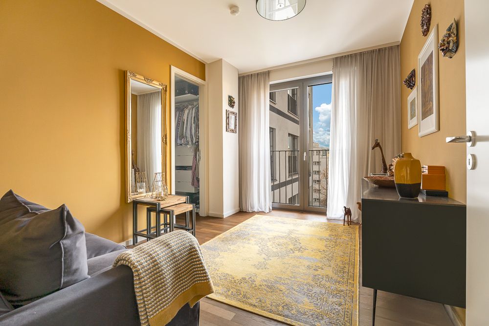 Helles Zimmer mit gelber Wand, Sofa, Spiegel, Teppich und Balkonfenster.