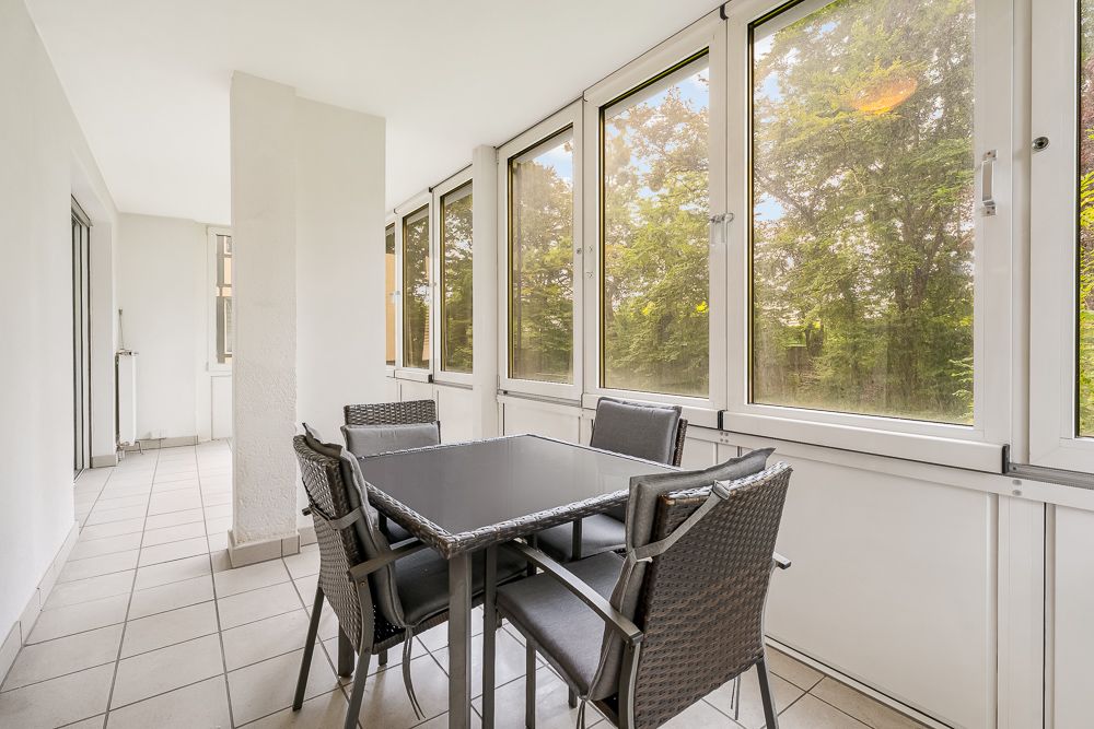Veranda mit großen Fenstern, Fliesenboden und einem schwarzen Tisch mit sechs Stühlen.