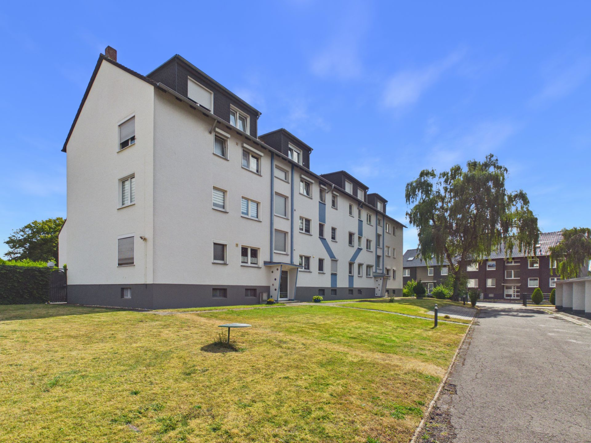 Mehrfamilienhaus mit weißer Fassade, grünem Rasen und Bäumen im Vordergrund.