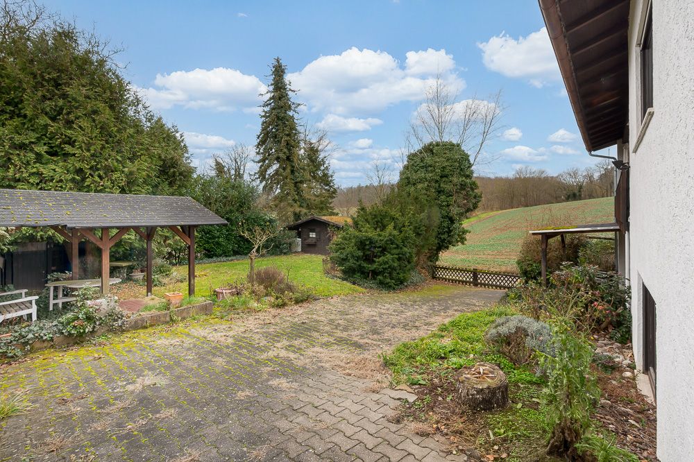 Gepflasterter Hof mit überdachtem Sitzplatz, Garten, Bäumen und angrenzender Wiese neben einem Wohnhaus.