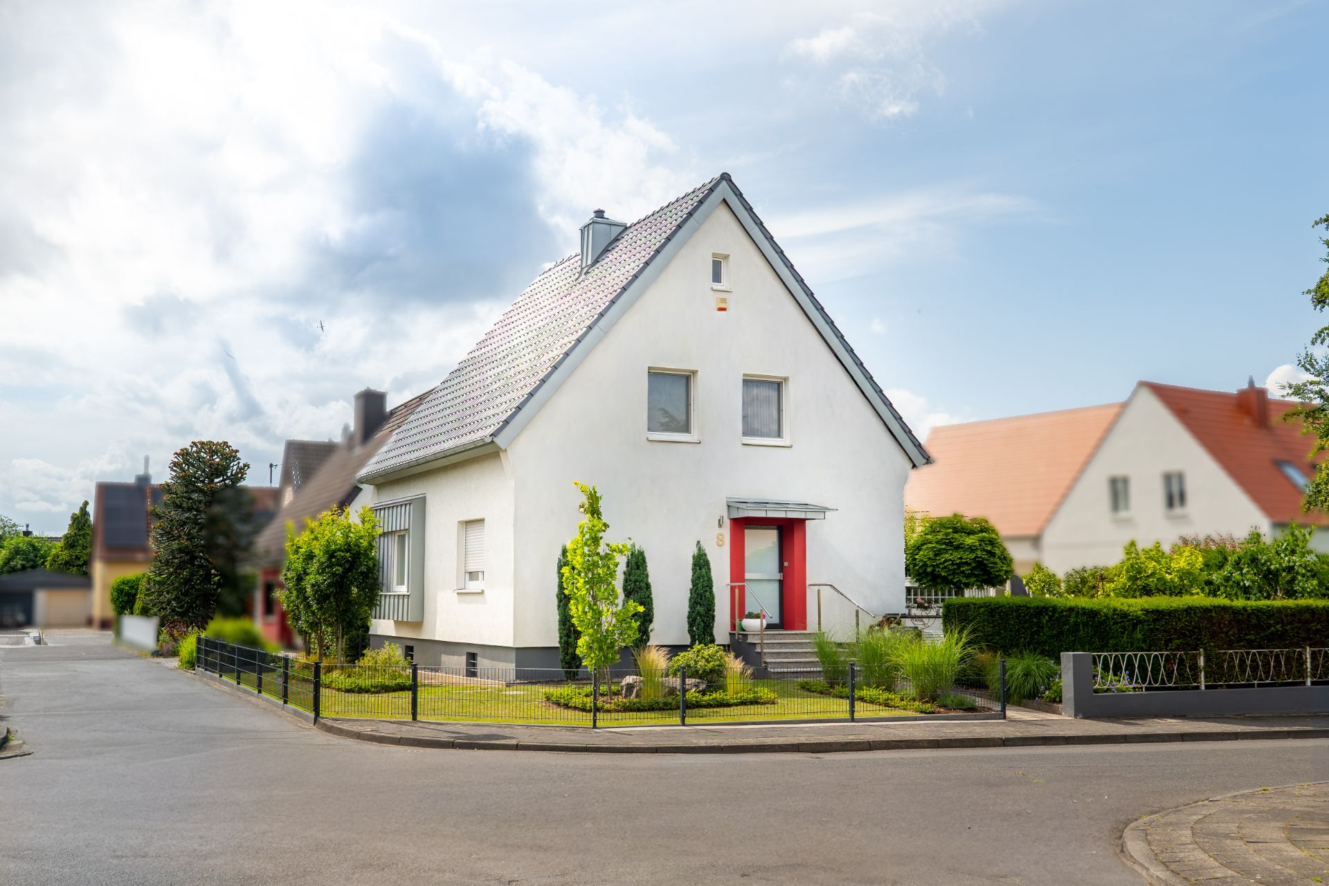 Weißes Einfamilienhaus mit rotem Eingang, Vorgarten und Zaun an einer Straßenecke.