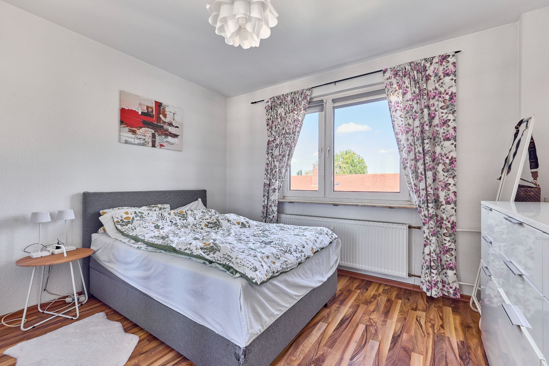 Helles Schlafzimmer mit Doppelbett, Fenster mit Blumenvorhängen und Holzboden.