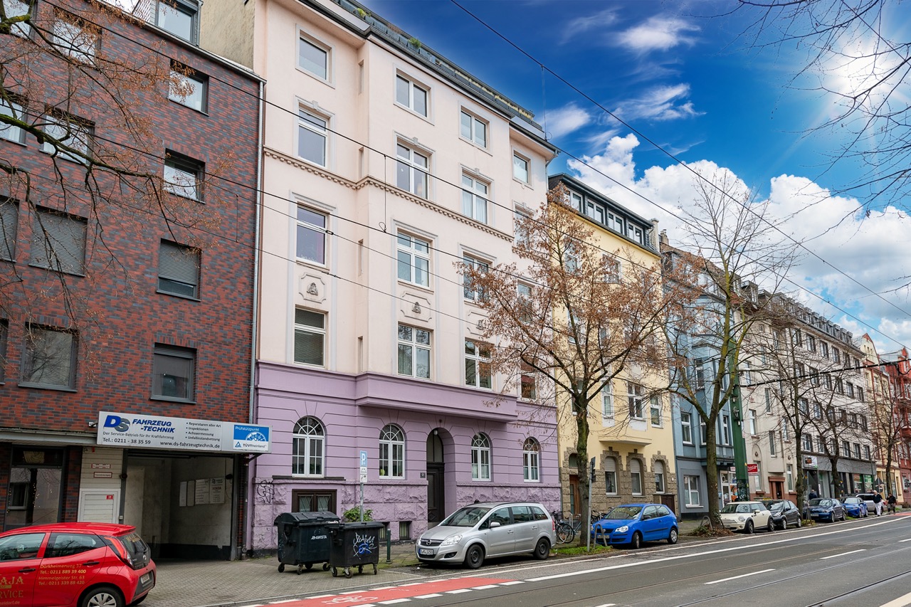 Mehrstöckiges Wohnhaus mit rosa Fassade, geparkten Autos und Bäumen an einer Straße.