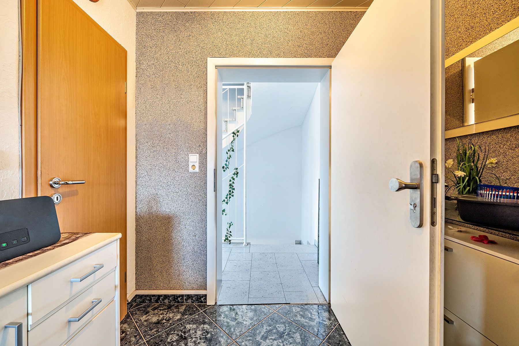Offene Tür zu einem Flur mit Treppe und weißen Wänden, daneben Schränke und ein Spiegel.