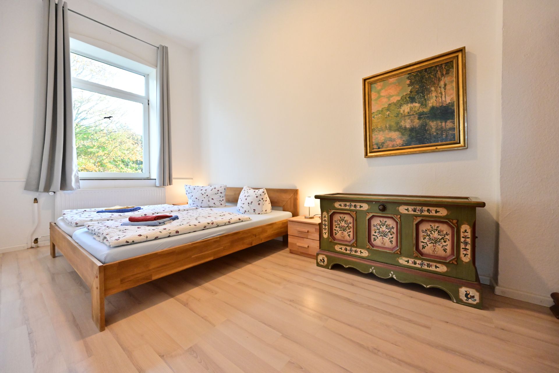 Helles Schlafzimmer mit Holzbett, Nachttisch, bemalter Kommode und großem Fenster.