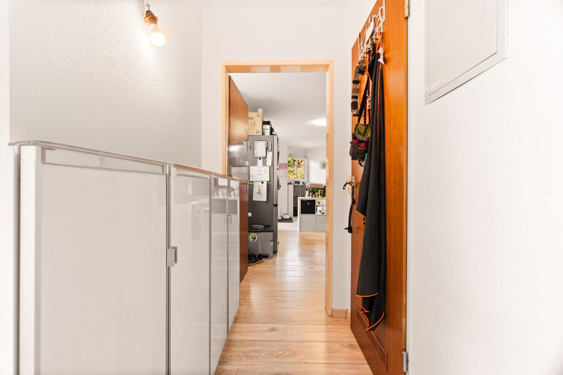 Flur mit Holzfußboden, Schrank und offener Tür, dahinter Küche und Wohnzimmer sichtbar.