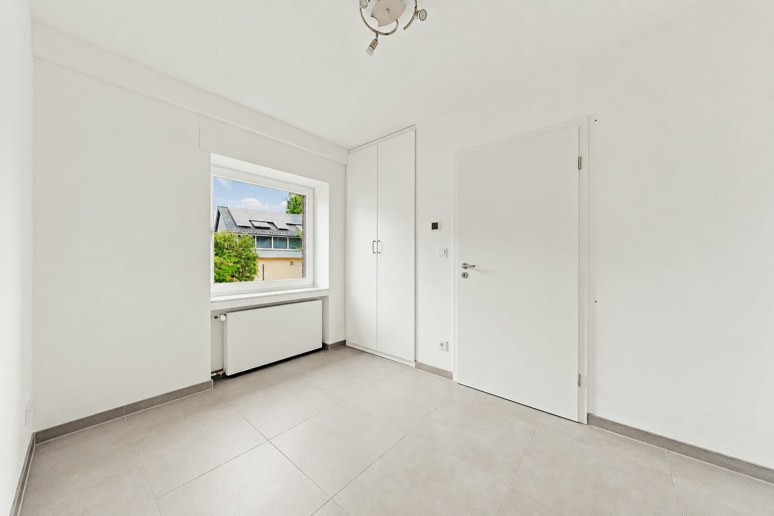 Helles, leeres Zimmer mit großem Fenster, weißer Tür und Einbauschrank.