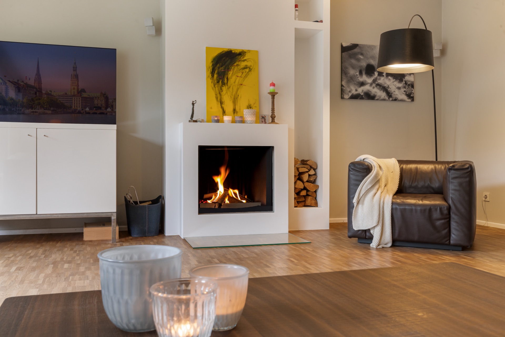 Wohnzimmer mit Kamin, Sessel, Fernseher, Kerzen auf Tisch und Holzvorrat.
