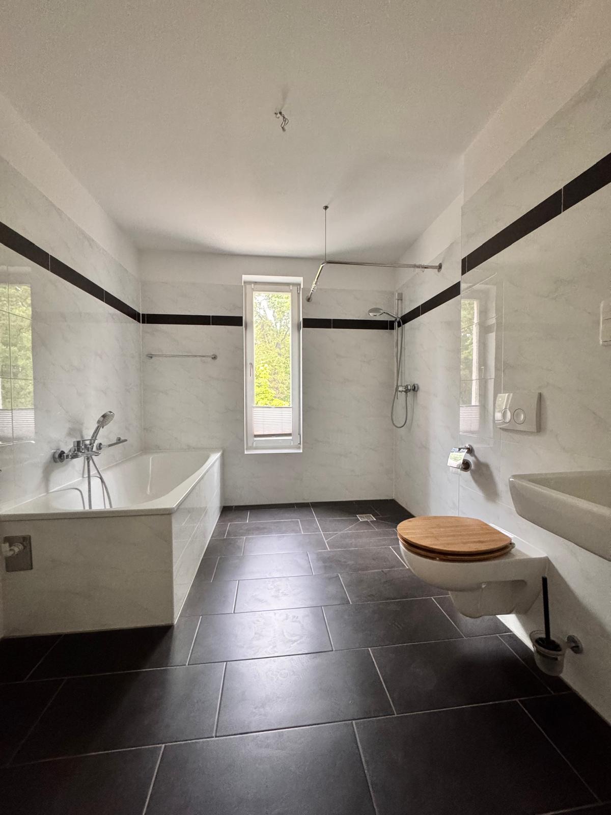 Modernes Badezimmer mit Badewanne, Dusche, WC und Fenster, dunkler Boden und weiße Fliesen.