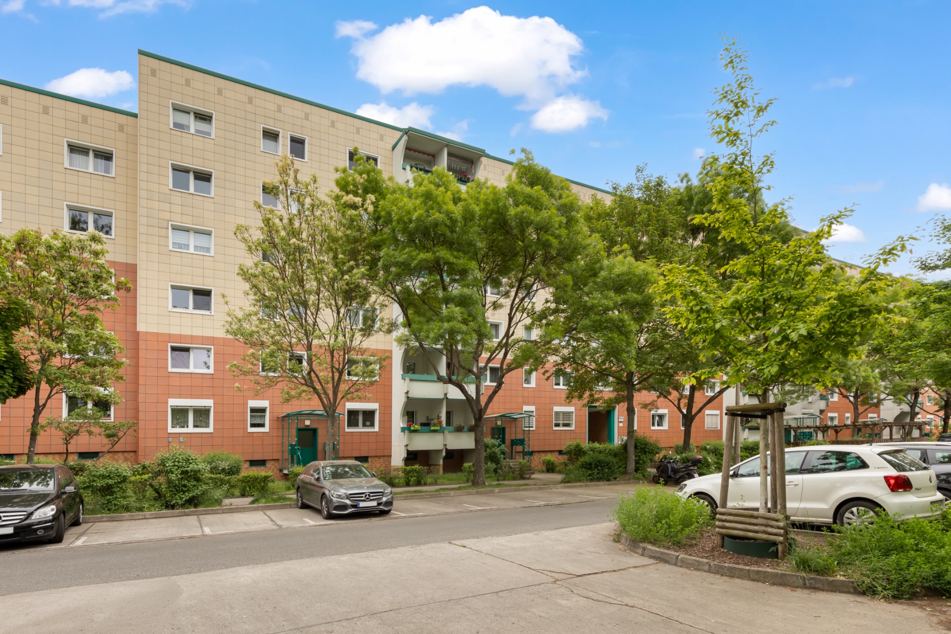 Mehrfamilienhaus mit Balkonen, davor Bäume, geparkte Autos und eine Straße an einem sonnigen Tag.