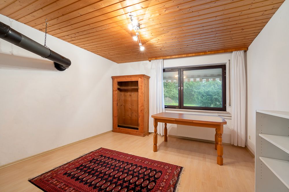 Helles Zimmer mit Holzdecke, Tisch, Teppich, Regal und großem Fenster mit Blick ins Grüne.