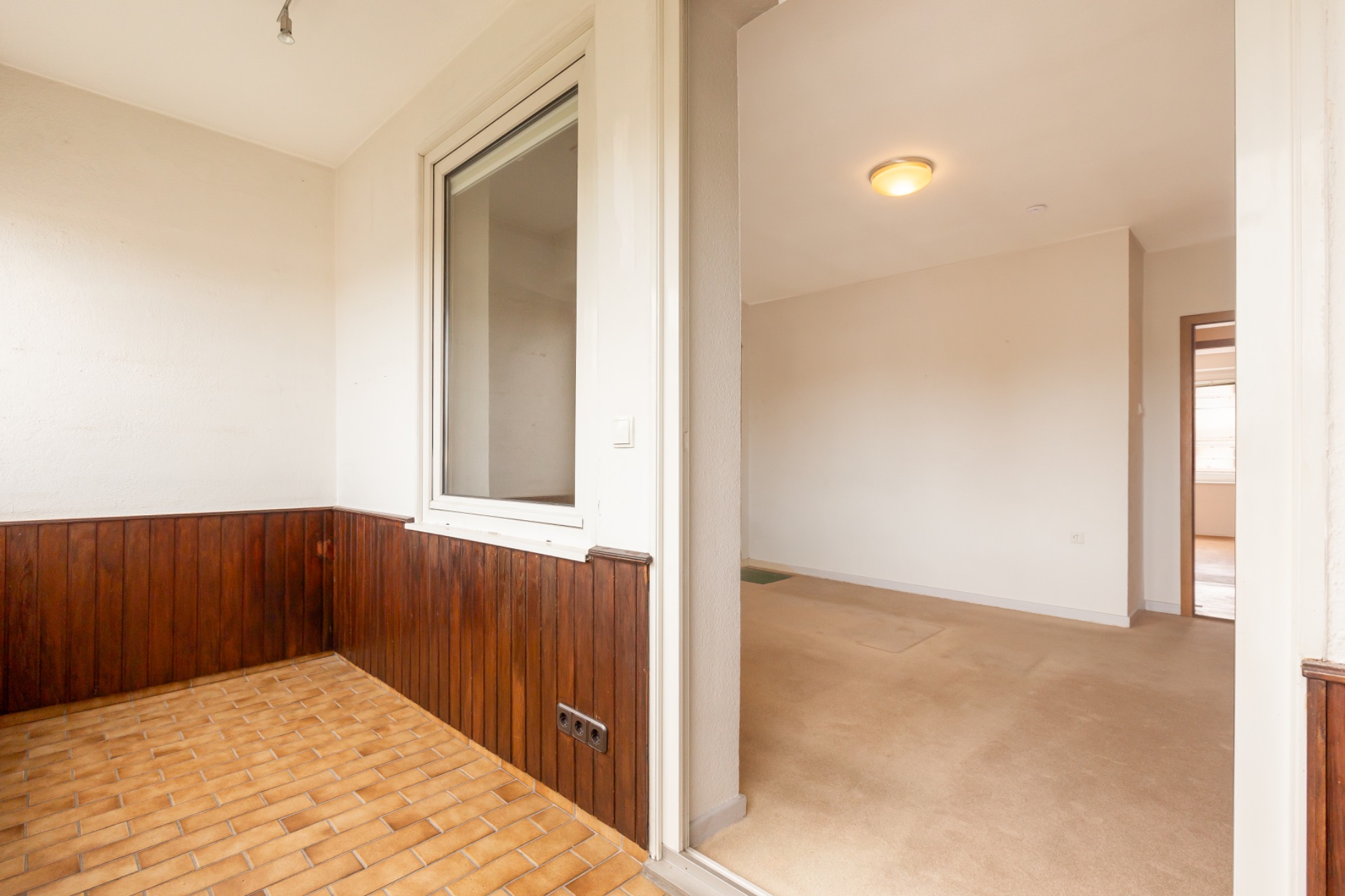 Innenraum mit gefliestem Boden, Holzverkleidung, Fenster und Durchgang zu einem Teppichzimmer.