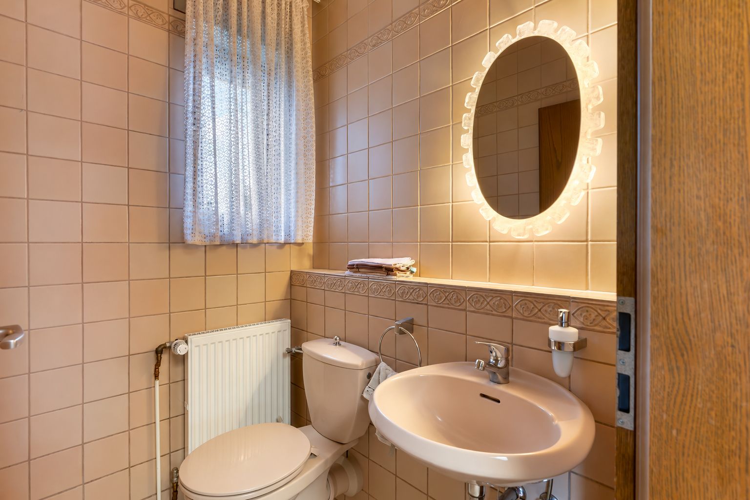 Kleines Badezimmer mit WC, Waschbecken, beleuchtetem Spiegel und Fenster mit Vorhang.