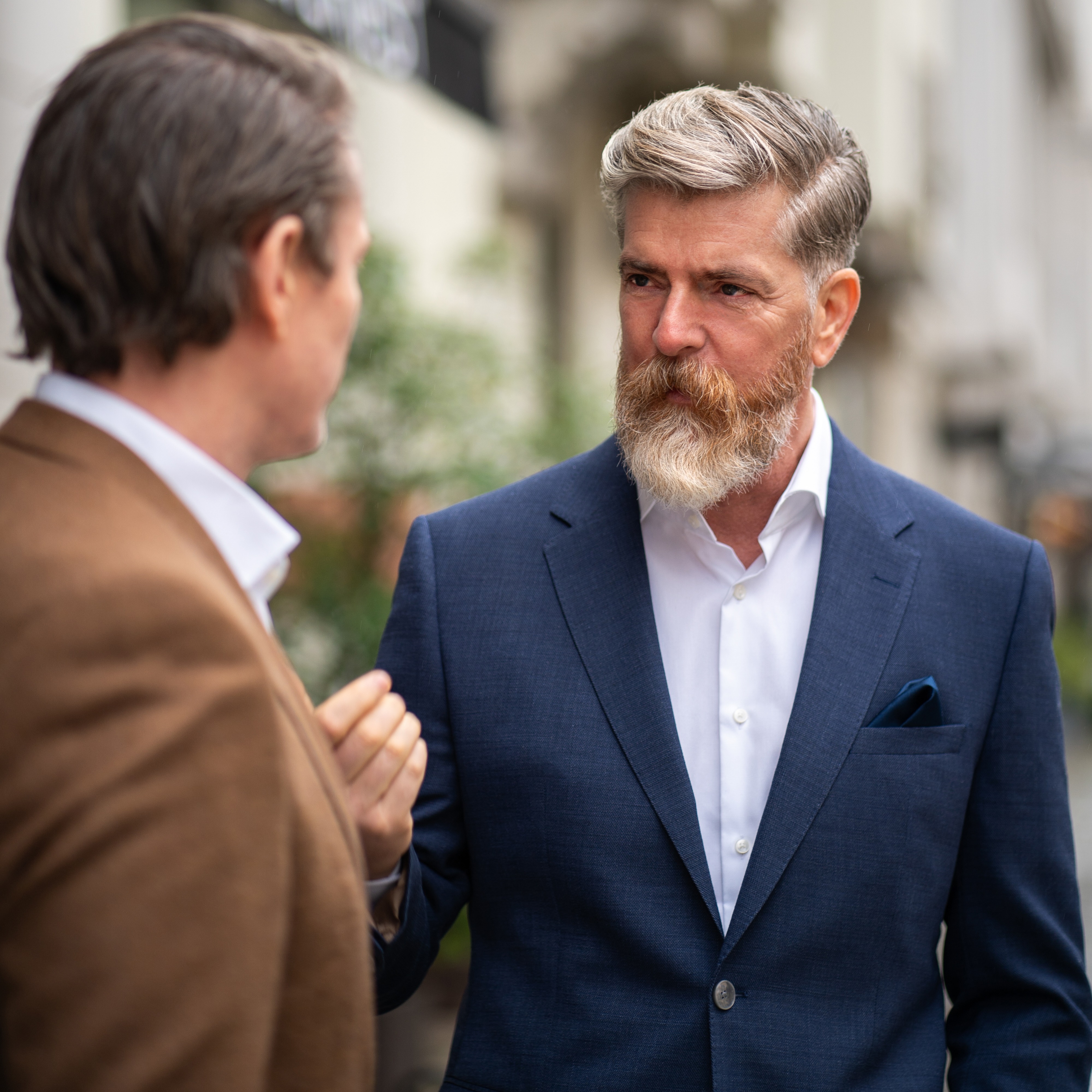 Zwei Männer im Anzug sprechen draußen vor einem Gebäude.