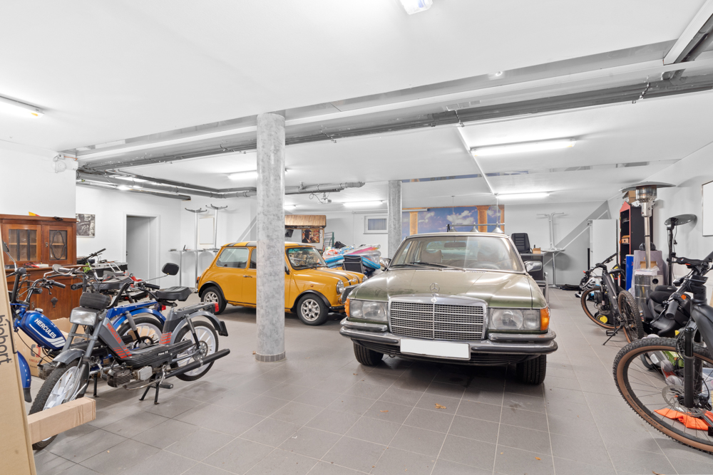 Große Garage mit mehreren Autos, Motorrädern und Fahrrädern auf grauem Fliesenboden.