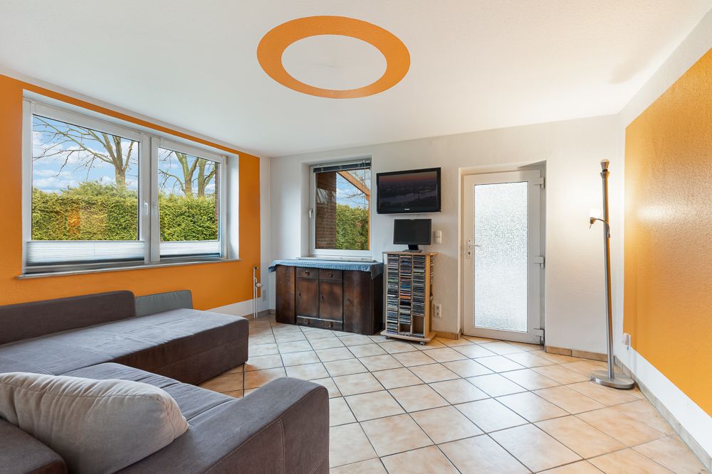 Helles Wohnzimmer mit orange gestrichenen Wänden, Ecksofa, Fliesenboden und großen Fenstern.