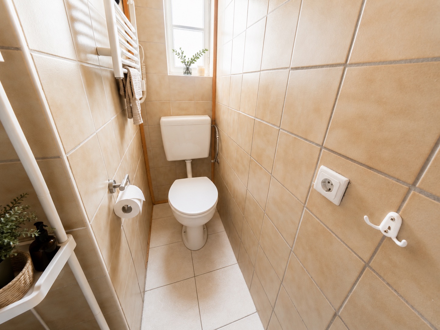 Schmaler gefliester Toilettenraum mit Toilette, Fenster und einigen Pflanzen.