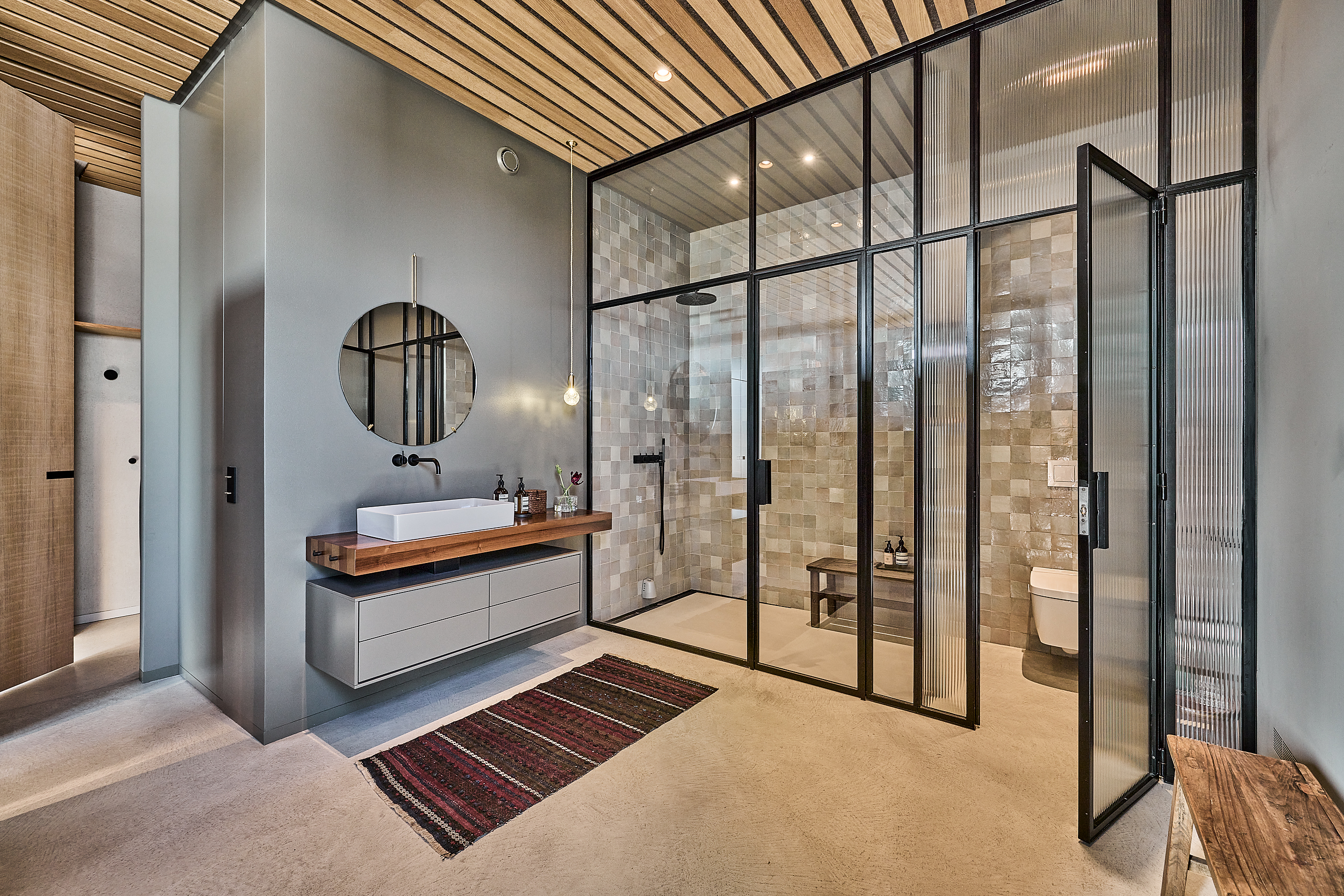 Modernes Badezimmer mit Waschbecken, rundem Spiegel, Glasduschwand und Holzbänken.
