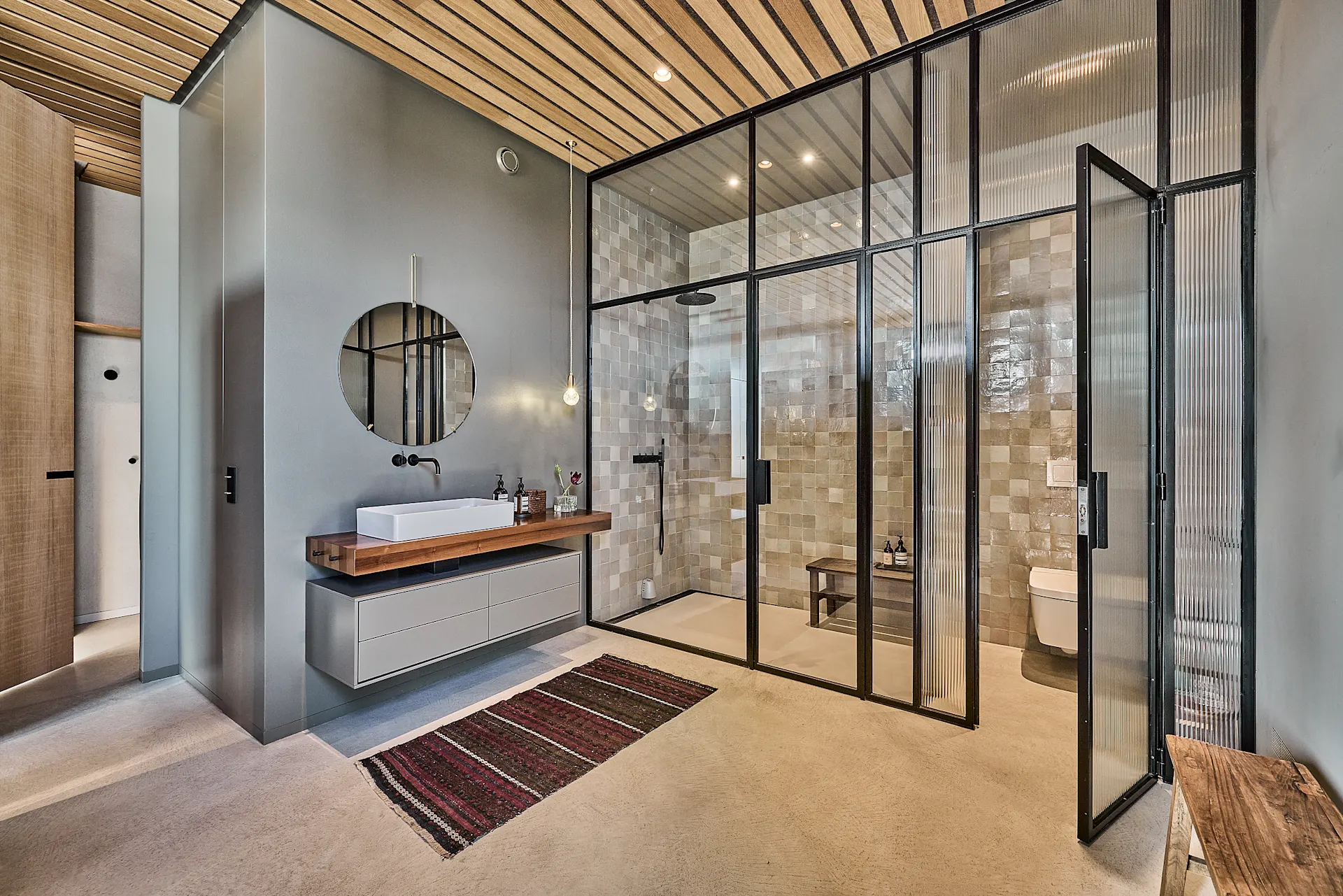 Modernes Badezimmer mit Waschbecken, rundem Spiegel, Glasduschwand und Holzbänken.