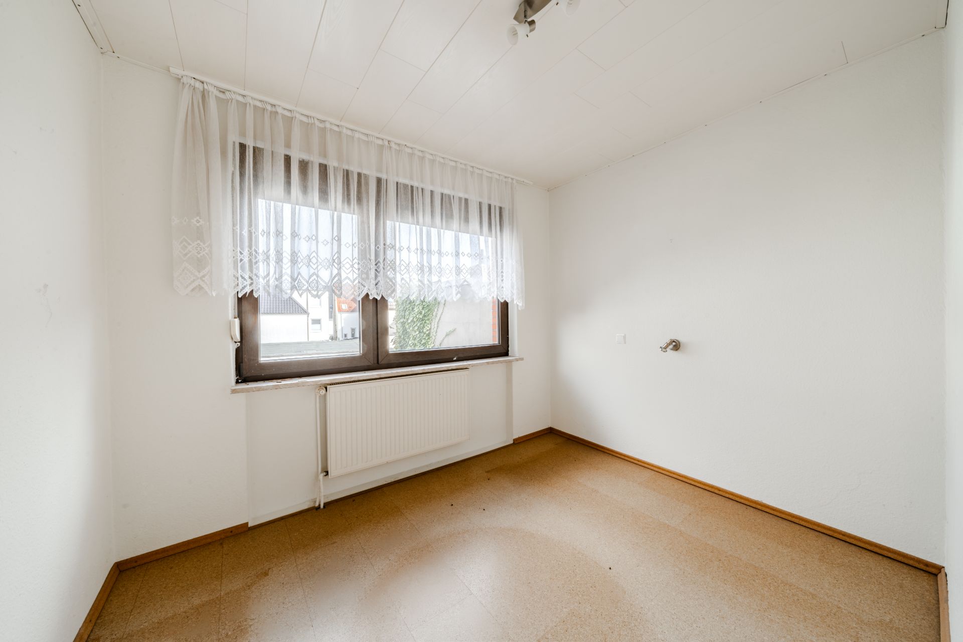 Leeres Zimmer mit großem Fenster, Heizkörper und weißer Gardine, weiße Wände und Holzboden.