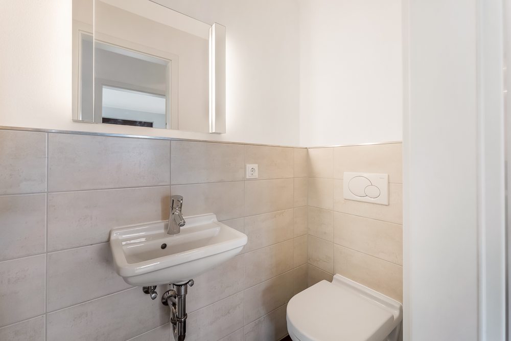 Kleines Bad mit Wandwaschbecken, WC, Spiegel und beiger Fliesenverkleidung.