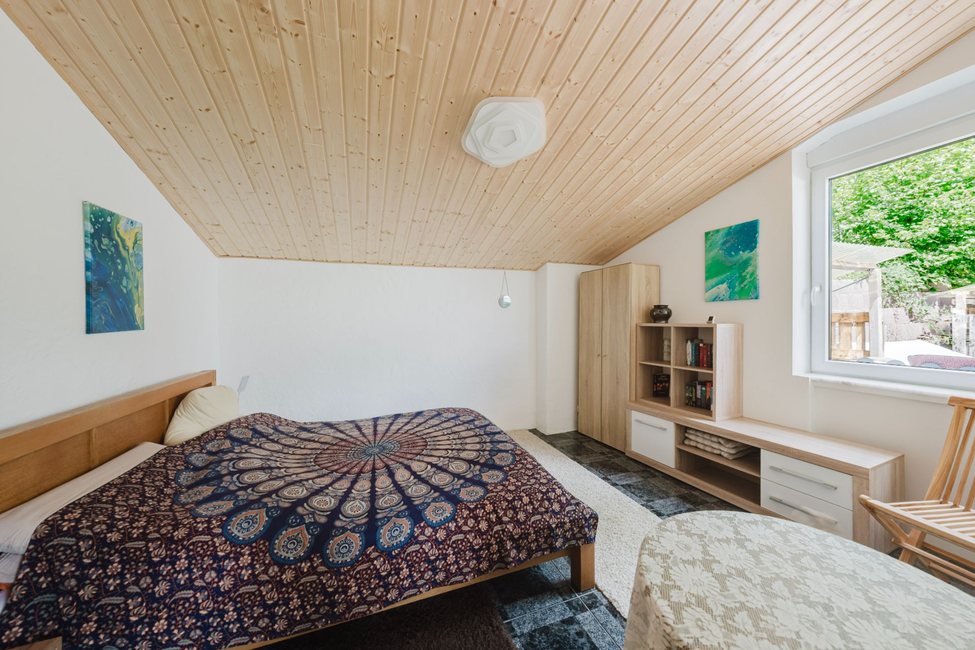 Schlafzimmer mit Holzbett, bunter Decke, Holzdecke, Schrank, Regal und großem Fenster.