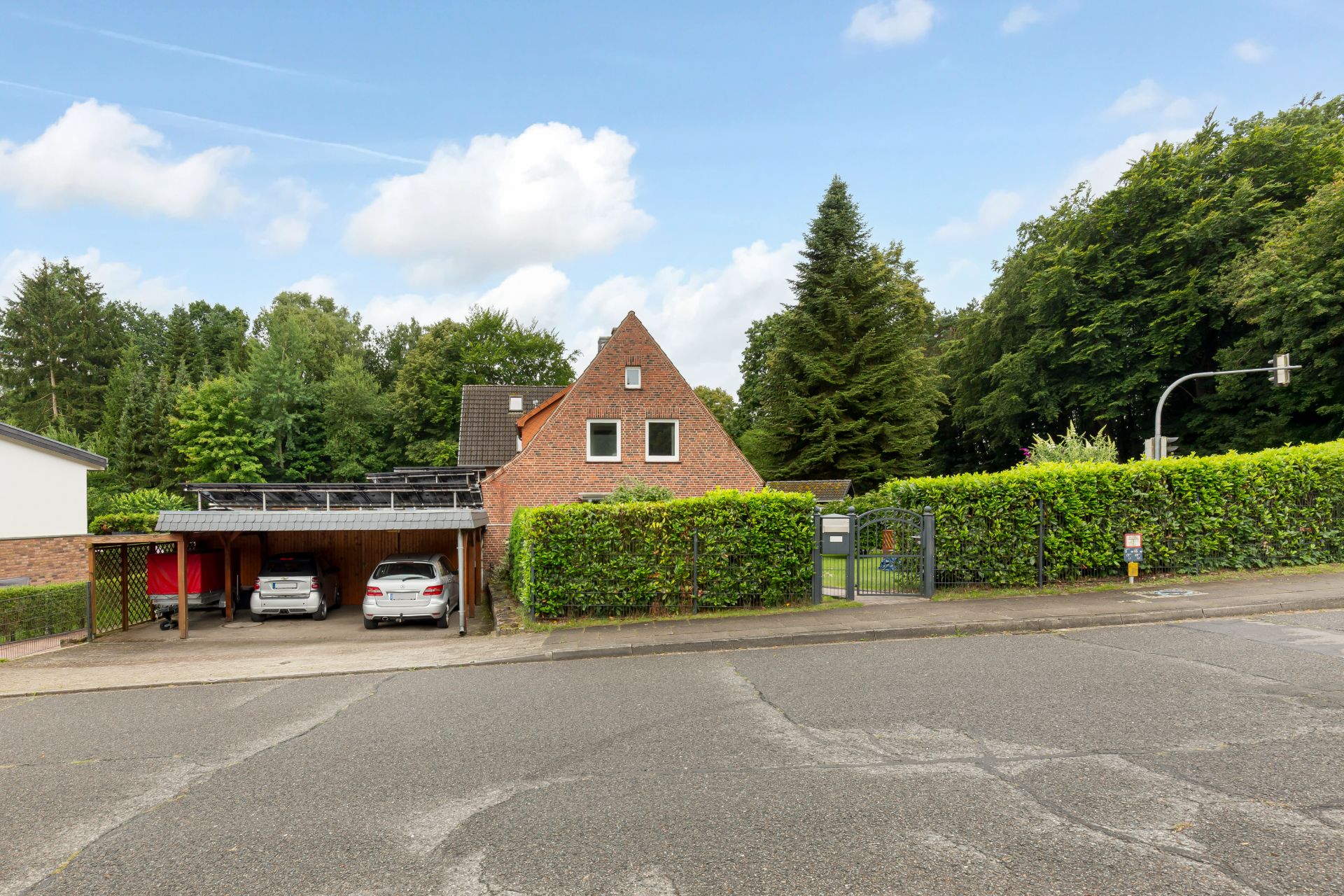 Backsteinwohnhaus mit Carport und zwei geparkten Autos, umgeben von Bäumen und Hecke.