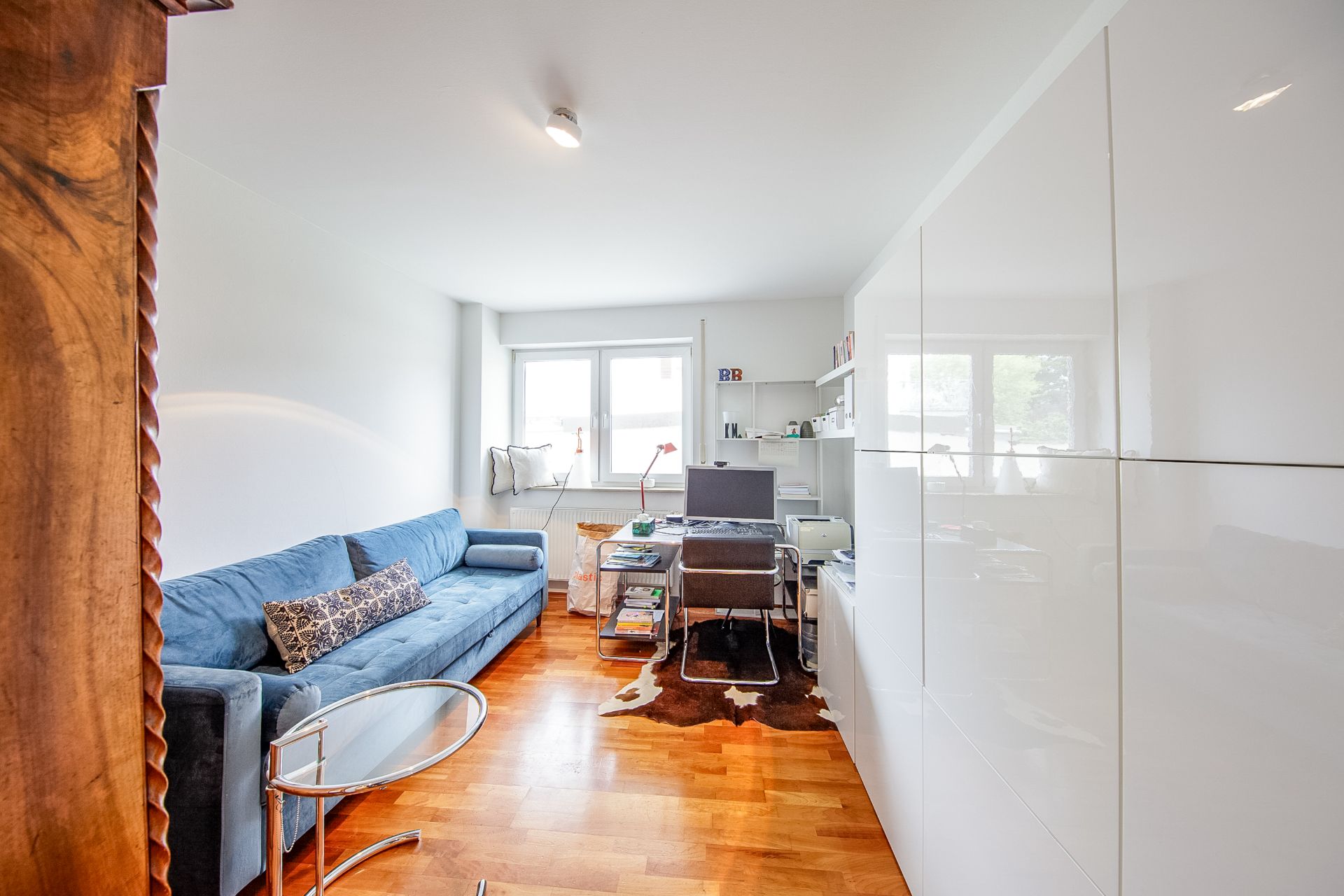 Helles Wohnzimmer mit blauem Sofa, Schreibtisch, Regal und großem weißen Schrank.